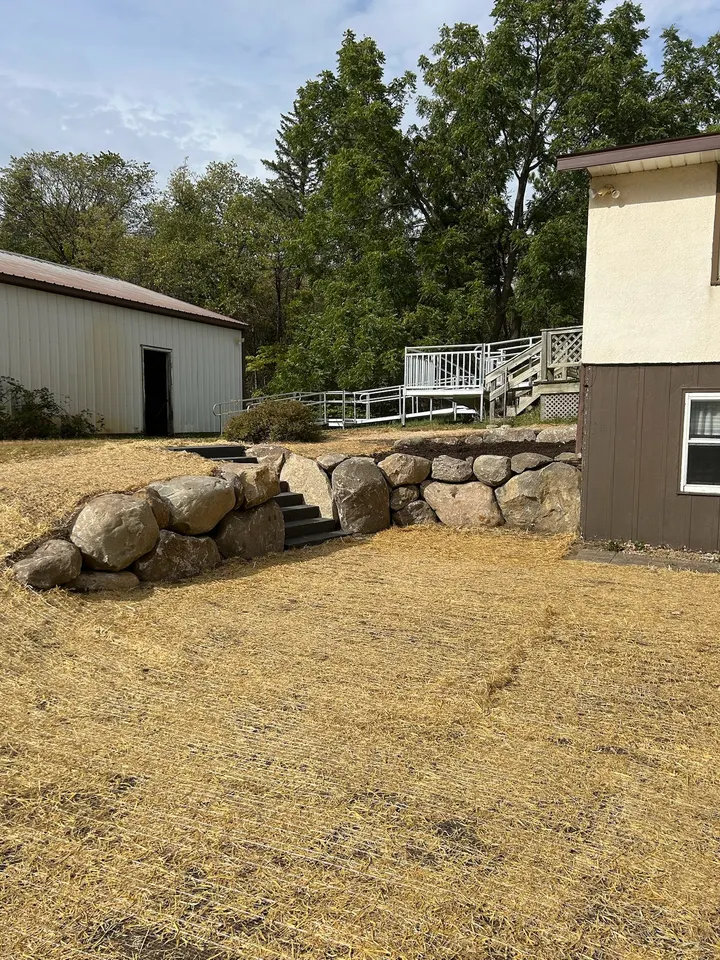 Boulder retaining wall with concrete steps set through it holding a freshly graded and straw-covered slope between a pole building and a house