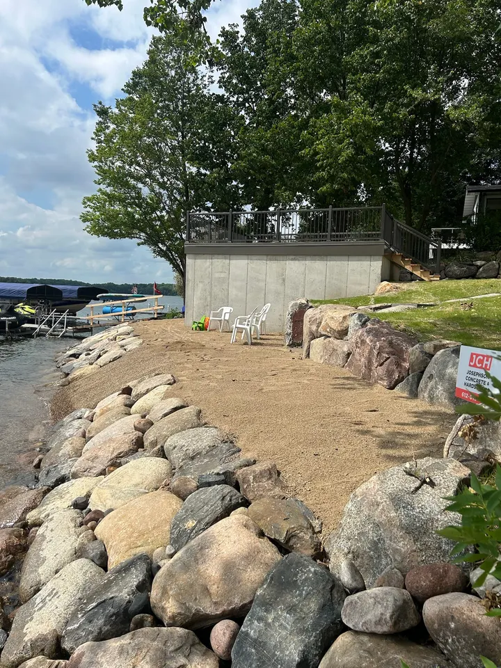 Boulder retaining wall stepping up a freshly graded lake bank with fieldstone riprap at the waterline and a house above