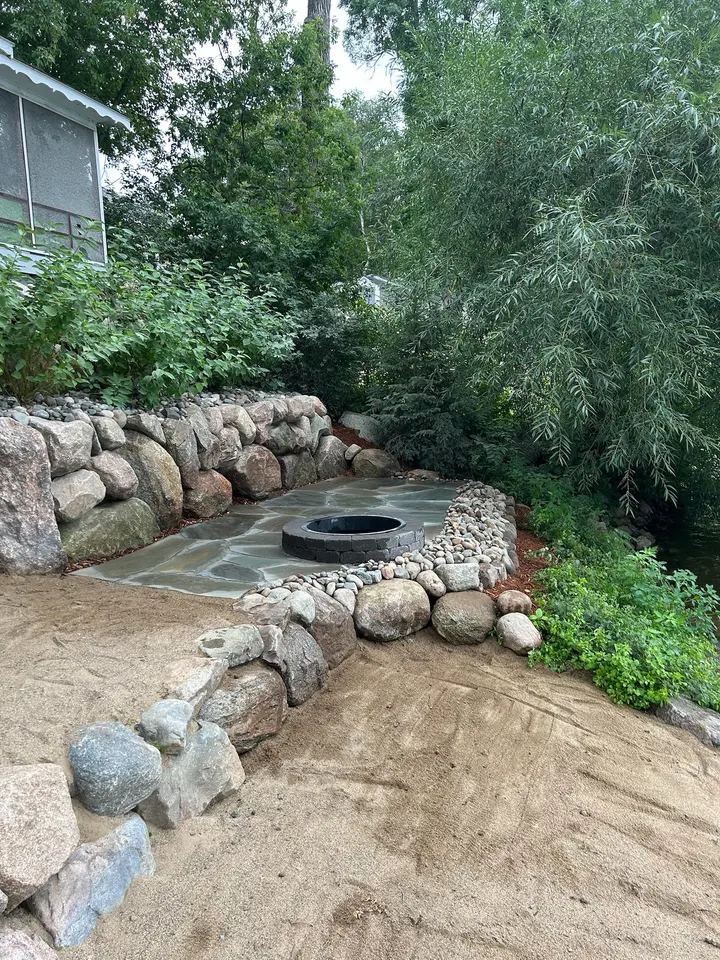 Boulder retaining wall wrapping around a paver patio with a fire pit, in a shaded backyard