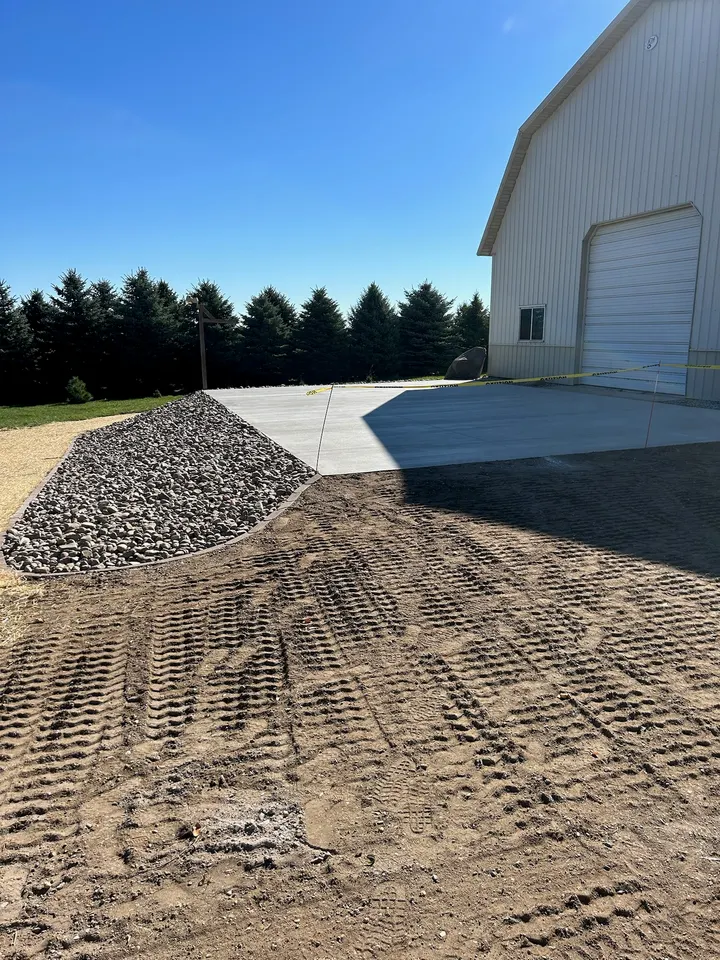 Compacted gravel base graded and ready for a concrete pour beside a white metal building, with a pile of gravel in the foreground