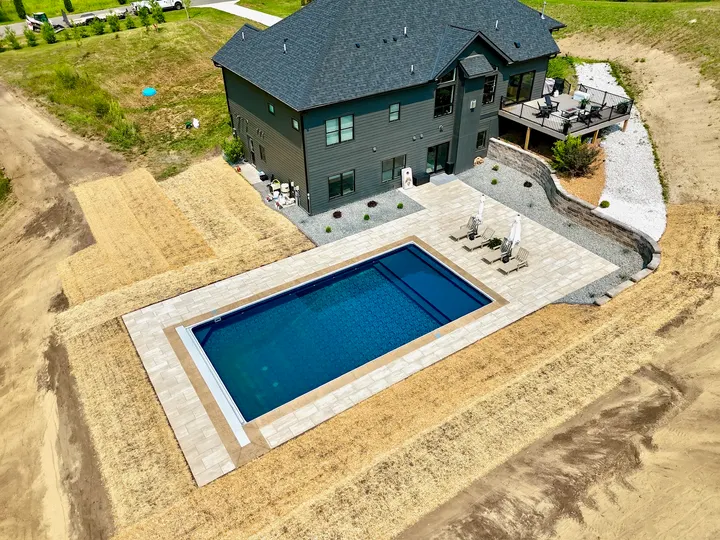 Drone view of a new rectangular pool with a concrete deck and paver border, surrounded by bare graded dirt beside a dark two-story home