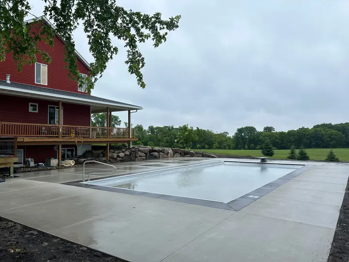 Wide concrete pool deck around a covered pool beside a red house with a deck, open field behind