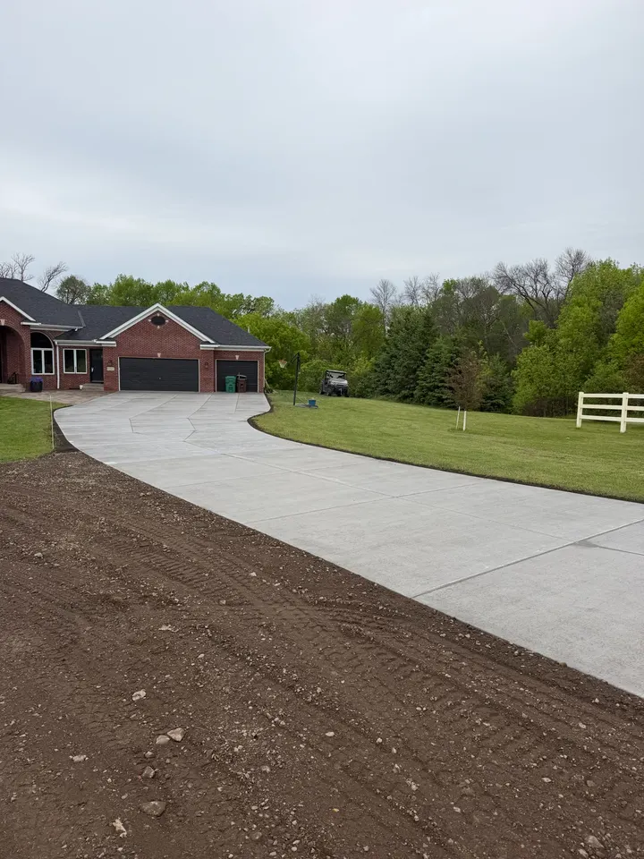 Long curved broom-finish concrete driveway sweeping toward a brick home in Hanover, MN, with fresh black dirt graded along both edges