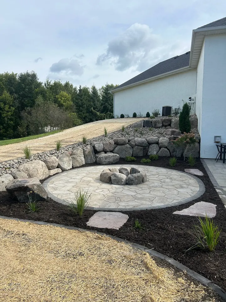 Round paver patio with a central fire pit, edged by a boulder retaining wall and fresh plantings