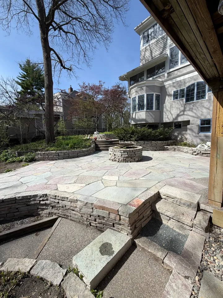 Flagstone patio with a round stone fire pit and low stone walls under a deck, with the house and trees behind