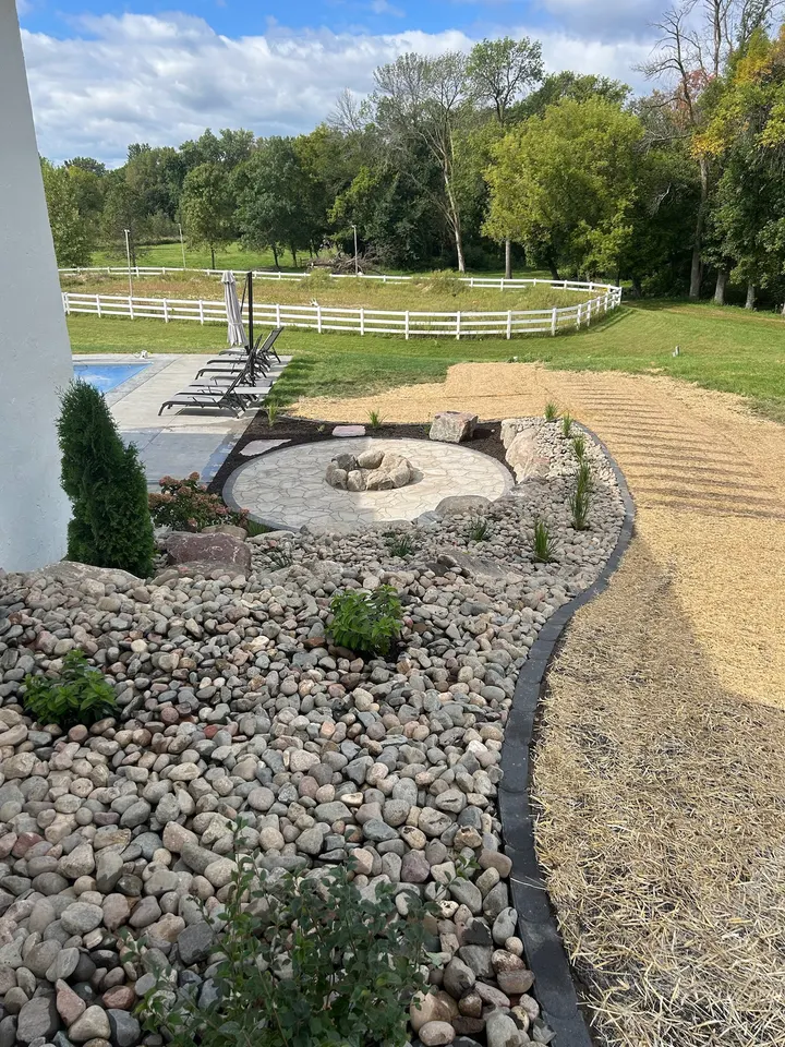 Curved walkway edged with river rock leading to a fire pit patio, with a white fence and pasture behind