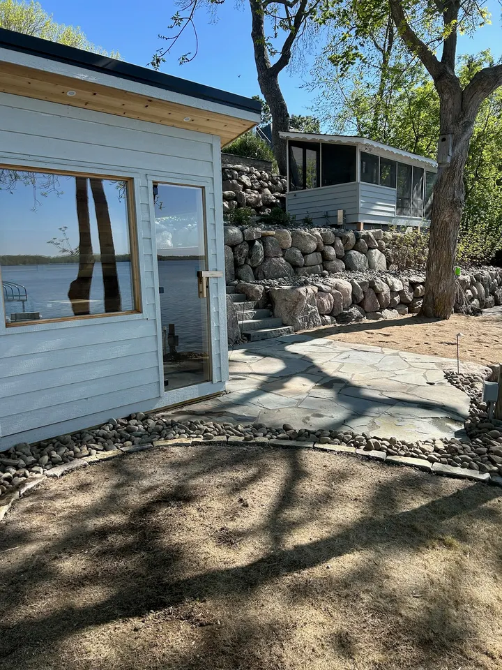 Boulder wall and stone patio beside a white lake shed under tall trees