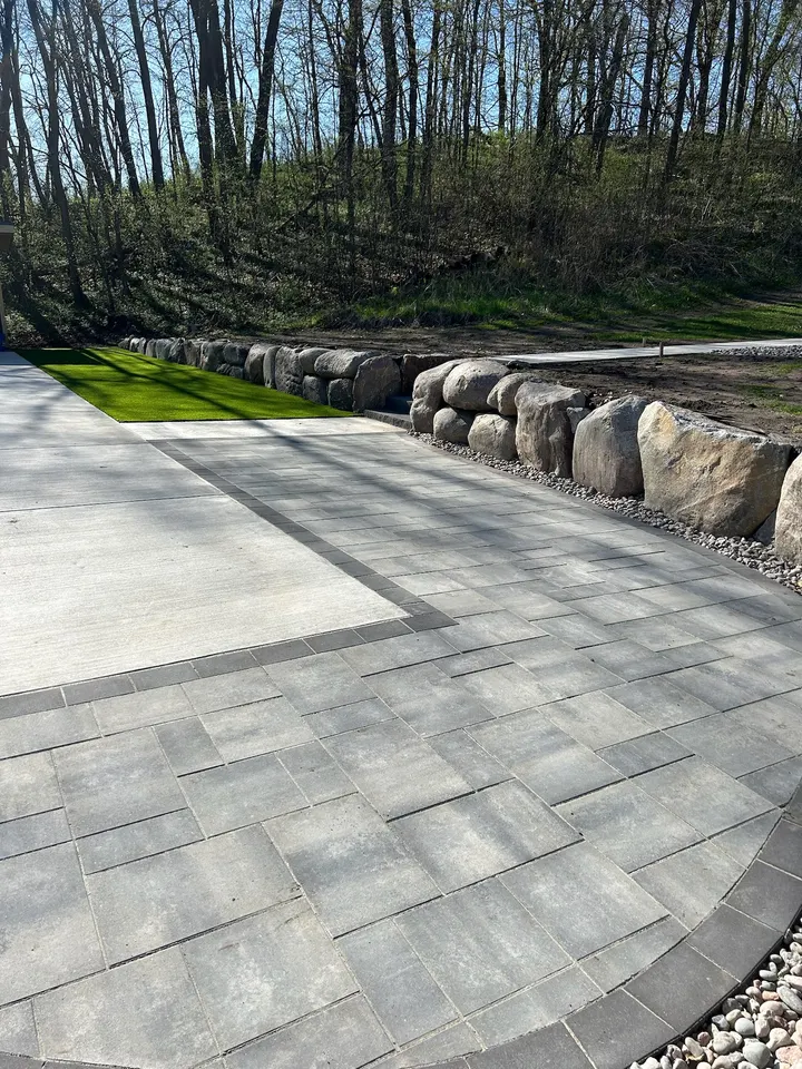 Large gray paver patio with a darker border, edged by a boulder retaining wall, trees behind