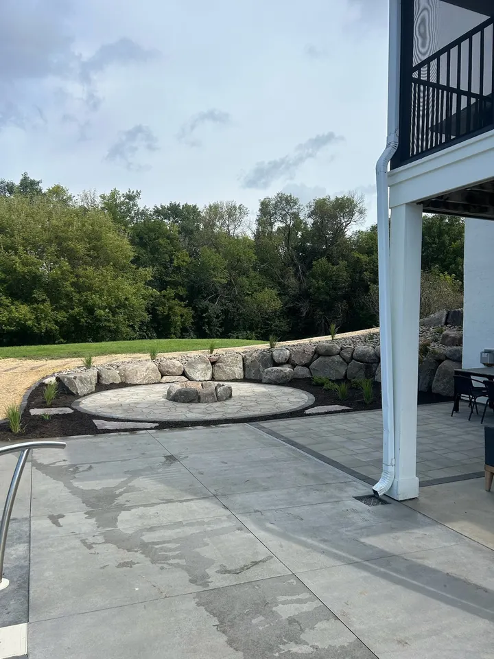 Round stone fire pit set into a concrete patio under a deck, boulder wall behind